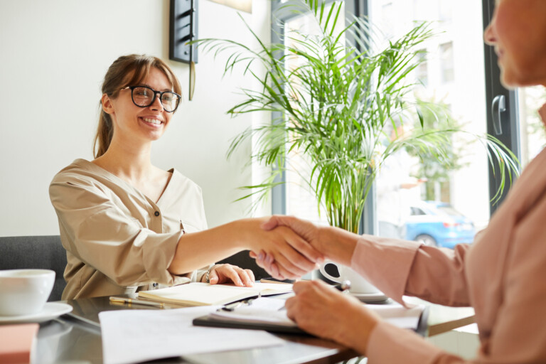 Young businesswoman in eyeglasses shaking hands to her partner and smiling while they working at the table at office